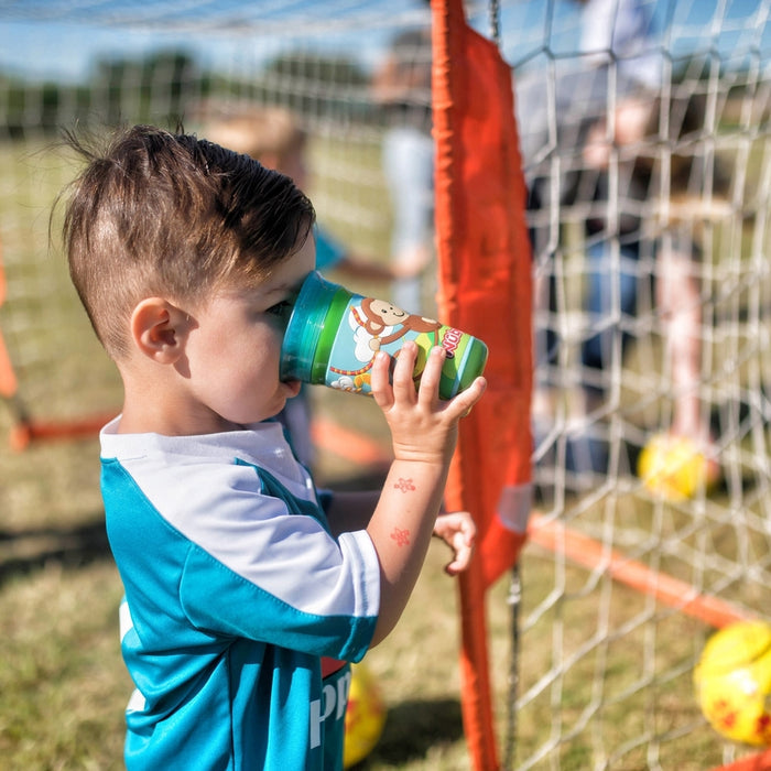 A young child in a blue soccer jersey drinks from a leak-resistant 360 Wonder Cup near a soccer goal, as yellow soccer balls and other kids playing can be seen in the background on a sunny day.