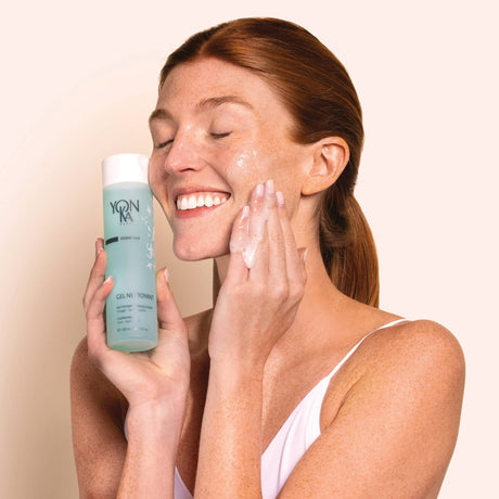 Woman with red hair smiling while applying facial cleanser to her cheek, holding a bottle of Gel Nettoyant Cleanser | Yon-Ka Paris skincare product in a minimal, light background setting.