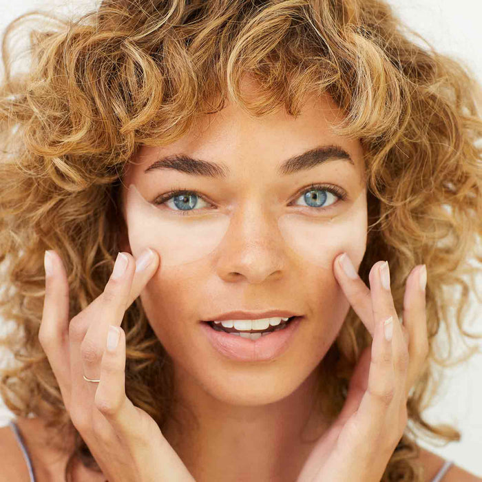 Close-up of a woman with curly hair applying the Under Eye Rescue Mask | Skin Authority under her eyes, demonstrating skincare for refreshed, hydrated skin.