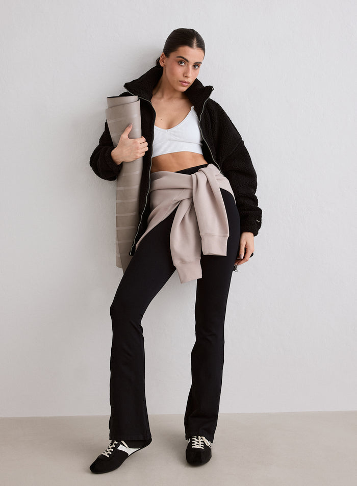 A woman wearing a black Pile Jacket, white sports bra and black leggings, holding a yoga mat [M-174cm]