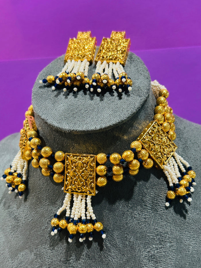 An ornate gold-toned jewelry set displayed on a gray mannequin bust against a purple background. The set includes a beaded necklace with rectangular filigree-style gold pendants, matching drop earrings, and white and navy blue bead tassels.