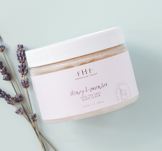 Jar of Honey Lavender Fine Sea Salt Body Scrub | Farmhouse Fresh next to sprigs of lavender on a light blue background, showcasing natural skincare and body care product.