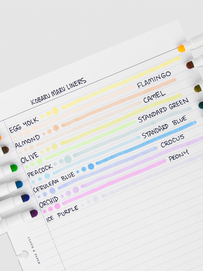 Pen test sheet showing examples of Egg Yolk, Flamingo, Almond, Camel, Olive, Standard Green, Peacock, Standard Blue, Cerulean Blue, Crocus, Orchid, Peony, Tourmaline, and Ice Purple ink. The pens are resting on the pen test sheet.