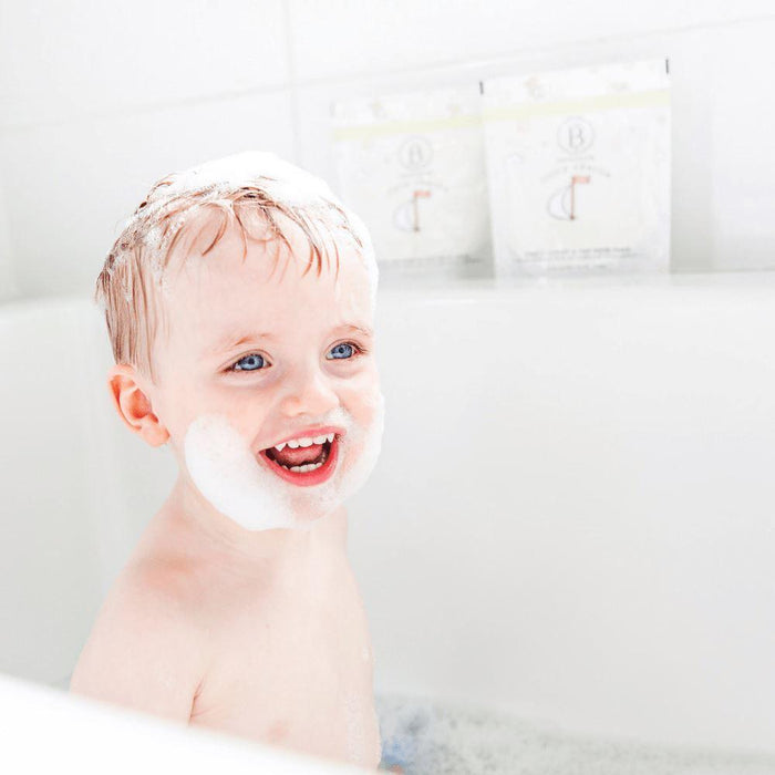 Smiling toddler enjoying a bubbly bath with white foam on head and chin, with two packets of little charlie™ Kids Bath Soak 250g | Bathorium in the background.