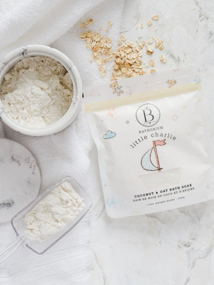 Package of little charlie™ Kids Bath Soak 250g | Bathorium next to a scoop and container filled with white bath soak powder on a white textured surface with scattered oat flakes.