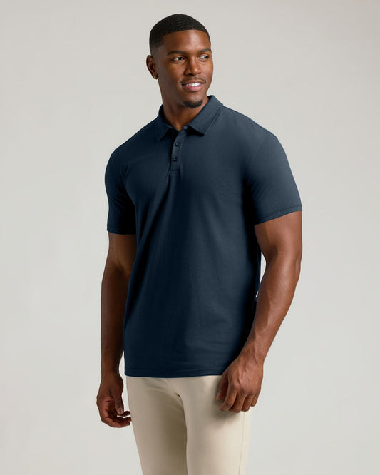 A man wearing a short-sleeve polo shirt in dark navy blue, paired with light beige pants, standing against a solid light gray studio background.
