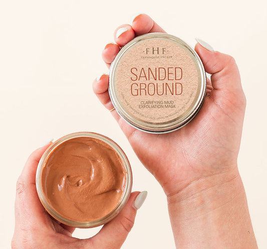 Hands holding an open jar and lid of Sanded Ground® Clarifying Mud Exfoliation Mask | Farmhouse Fresh, showing the smooth brown exfoliating mask inside the jar.