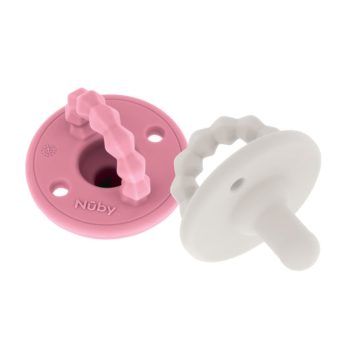 Two Nuby Silicone Softees Pacifier & Teether (2 Pack), one pink and one white, each with a round shield and textured handle. The pink pacifier features the Nuby brand name. Both are displayed on a white background.