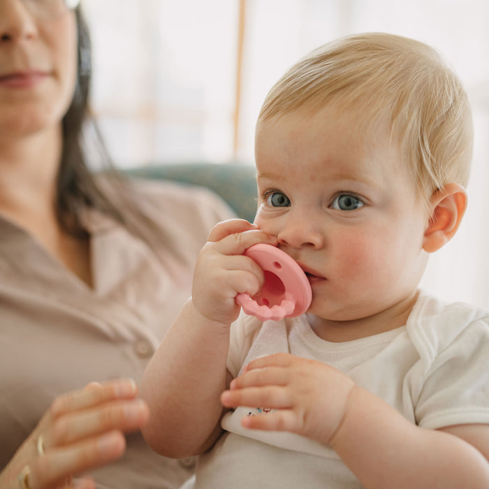 A baby with blonde hair sits on an adult’s lap, chewing on a pink Nuby Silicone Softees Pacifier & Teether (2 Pack) for soothing, while the adult’s face is partially visible in the background.