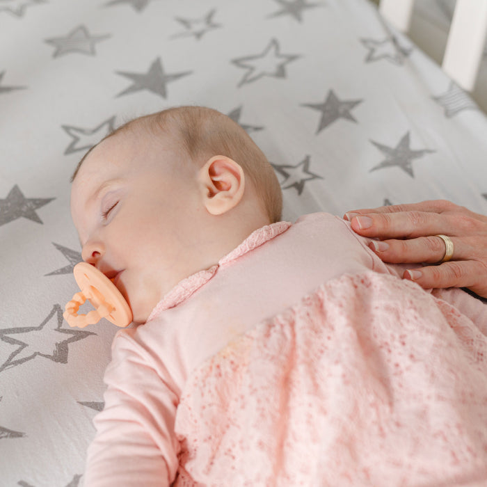 A baby in pink sleeps on star-patterned sheets with a Nuby Silicone Softees Pacifier & Teether (2 Pack) in their mouth, while an adult hand gently rests on the baby’s side, creating a soothing moment.