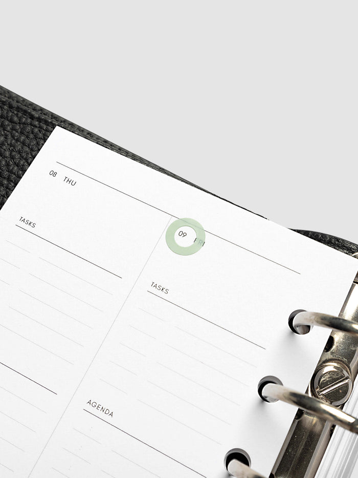 A close-up of a weekly planner page inside a black textured ring binder, showing lined sections for tasks and agenda with a small green sticker highlighting the date.