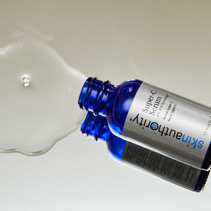 Blue glass bottle of Super-C Serum | Skin Authority laying on reflective surface with clear serum spilled beside it, highlighting skincare and wellness.