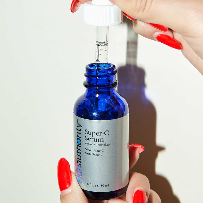 Hand holding a blue glass dropper bottle of Super-C Serum | Skin Authority with bright red polished nails, applying skincare serum with a dropper against a white background.