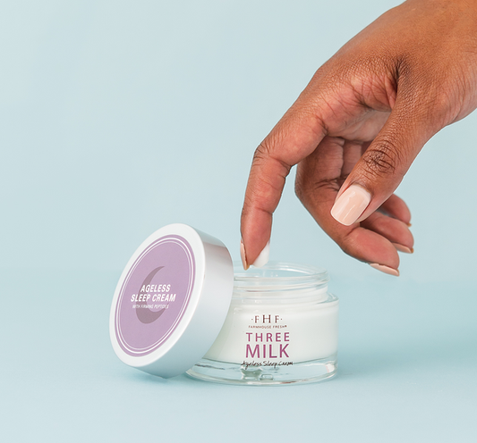A hand dipping a finger into a jar of Three Milk Ageless Sleep Cream with Peptides | Farmhouse Fresh against a light blue background.