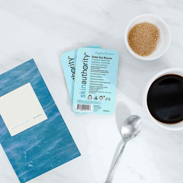 Two light blue packets of Under Eye Rescue Mask | Skin Authority placed on a white marble surface next to a cup of black coffee with a spoon and a small bowl of brown sugar beside a blue book titled Recipes.