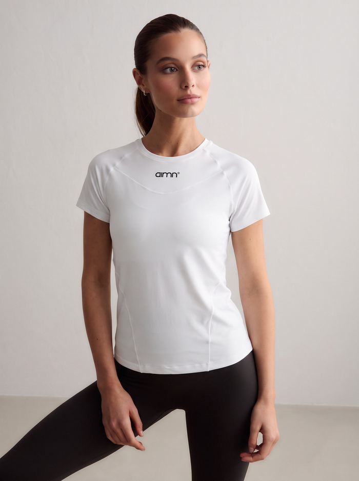 A model wearing the white Soft Basic Short Sleeve with a printed aim'n logo, paired with black tights [S-174 cm]