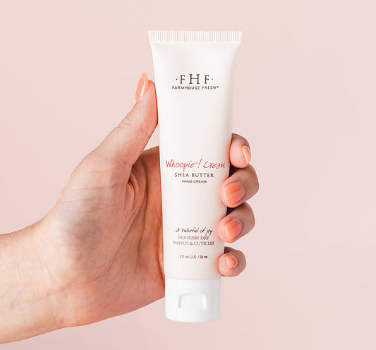 Hand holding a tube of Whoopie® Cream Shea Butter Hand Cream | Farmhouse Fresh against a soft pink background, showcasing skincare product for nourishing dry hands and cuticles.