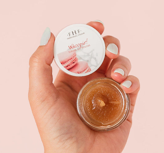 Hand holding an open jar of Whoopie® Lip Polish | Farmhouse Fresh with sugar lip polish visible inside against a soft pink background.